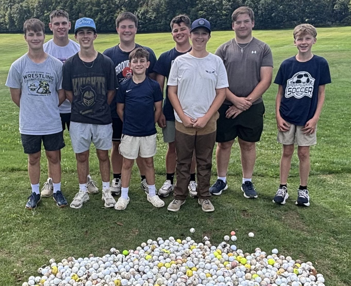 Nine members of the golf team stand behind a pile of golf balls out on the course.